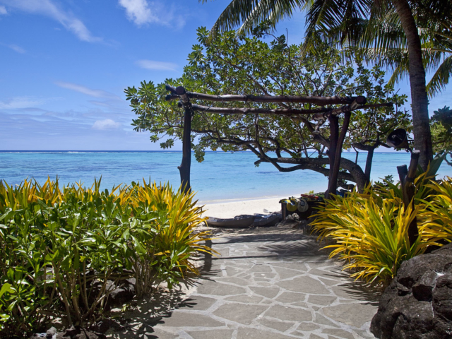 Hard-wood-archway permanently built as entrance to and from the beach showcasing the green and yellow flowers neatly planted along the pavement to compliment the tempting blue lagoon in the background