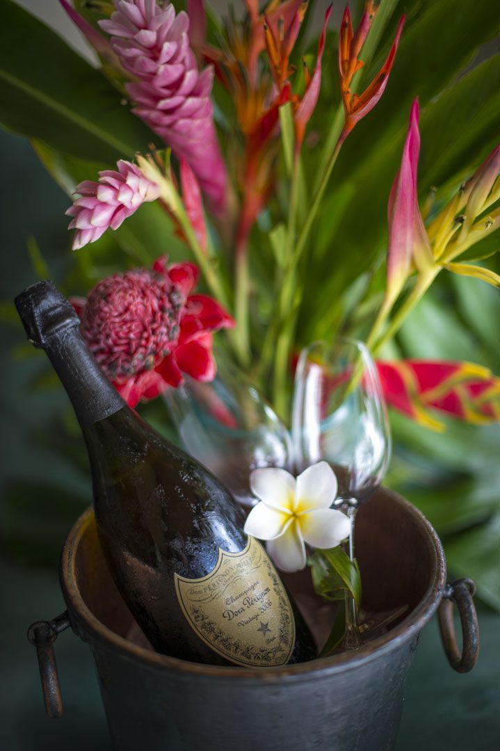 A chilled bottle of Champagne with flutes in a bucket of ice, set up next to a tropical floral arrangement