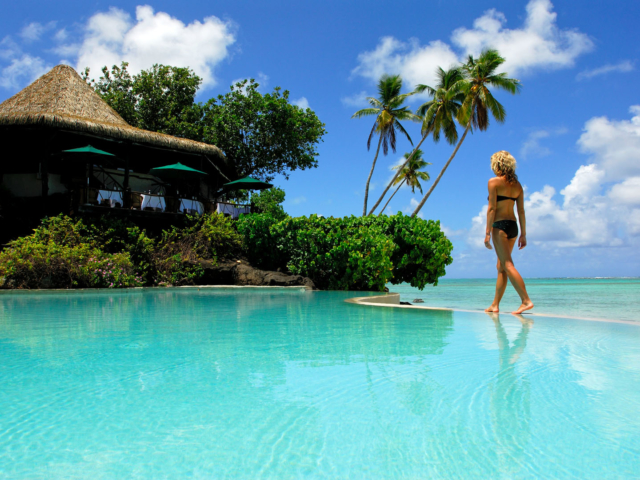 A resort guest walks along the edges of the swimming pool on a clear bright day featuring a stunning aqua-coloured pool complimenting the shades of the lagoon in the background