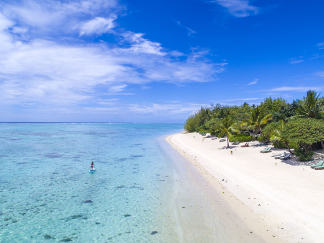 Aerial image of a Resort guest paddle-boarding in front of the Resort, parallel to the lined up sun loungers neatly placed in pairs on a long white stretched sandy beach on a beautiful clear day