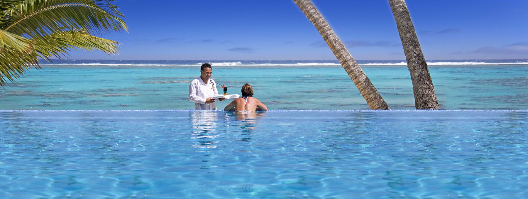 A woman accepting a cocktail from a waiter while admiring the beach views from the pool