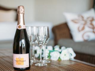 A bottle of champagne placed on a table with four glasses and white roses in the background as a sign of celebration