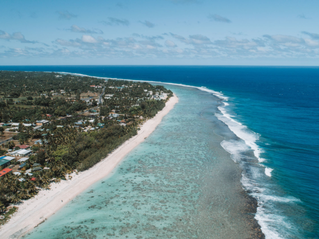 An aerial partial image of Rarotonga Island showing off the splashing of waves on the reef and the various blue shades of the Rarotonga waters