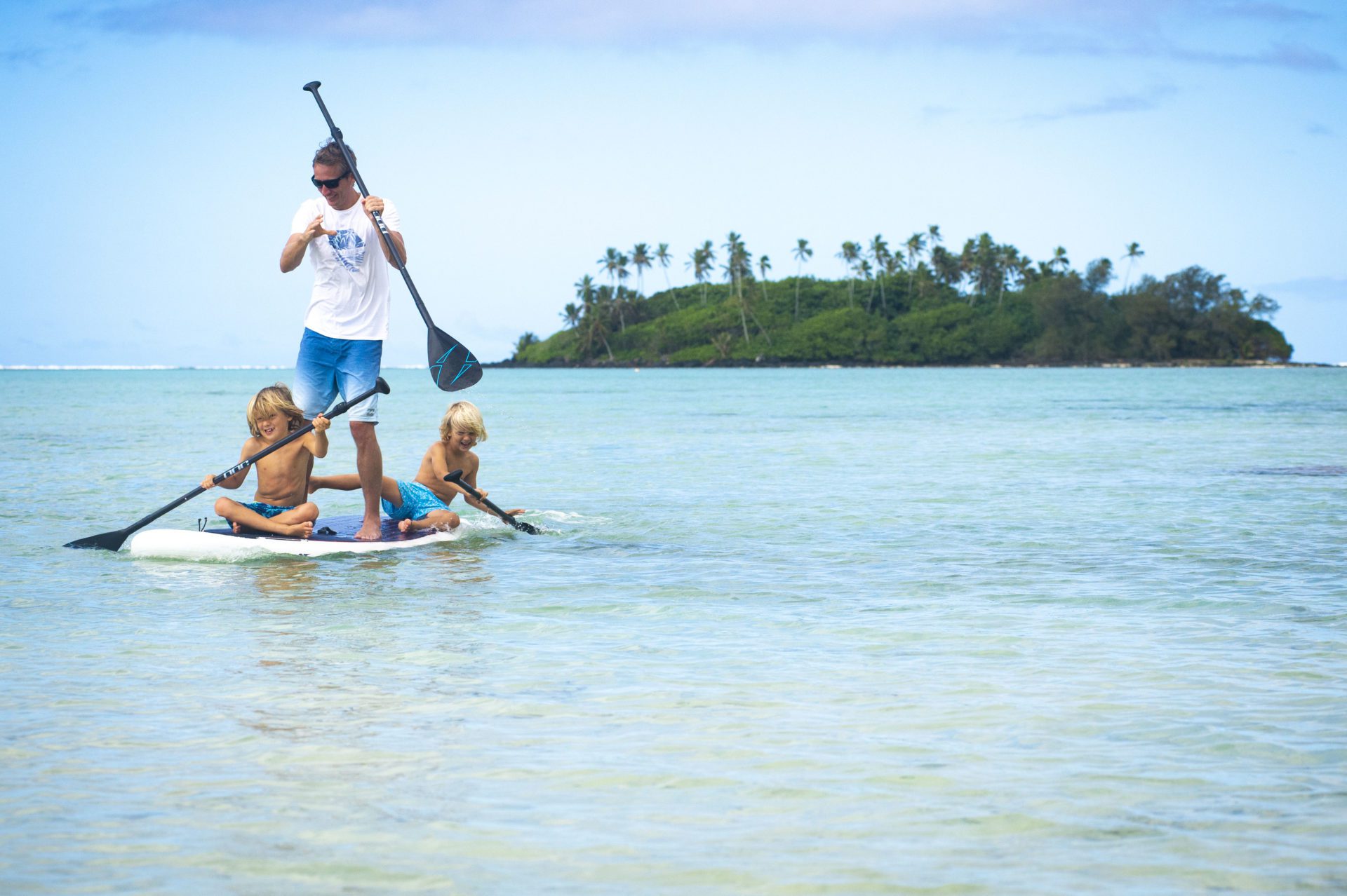 A beautiful image of father and two sons bonding in the lagoon sharing a stand-up paddleboard and smilingly paddles through the waters of Muri