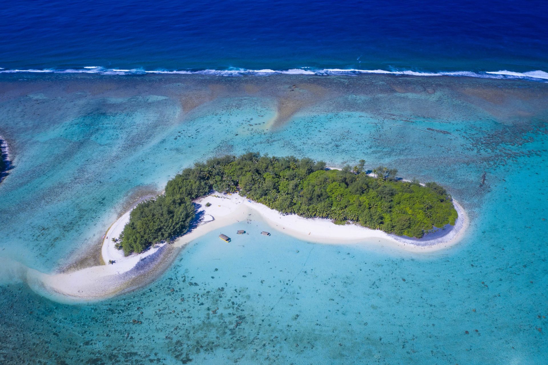 A spectacular aerial image of Motu Koromiri, a secluded private island set amidst the clear blue lagoon showcasing the splashing of waves towards the outer reef