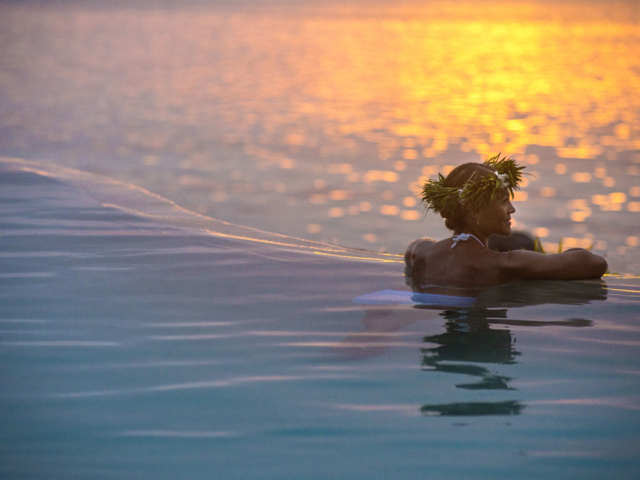 Guest in the pool, leans towards the edge to enjoy the serene sunset reflection on the lagoon as it goes down the horizon giving its last shade of yellow and orange