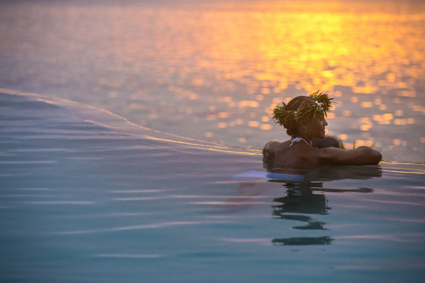 Guest in the pool, leans towards the edge to enjoy the serene sunset reflection on the lagoon as it goes down the horizon giving its last shade of yellow and orange