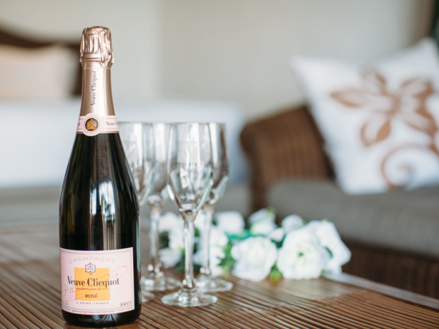 A bottle of champagne placed on a table with four glasses and white roses in the background as a sign of celebration