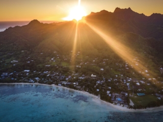 A stunning aerial image of the South-Eastern side of Rarotonga Island capturing the spread out view of the Muri lagoon view up to the mountain peaks leaving the glorious sunset as a background