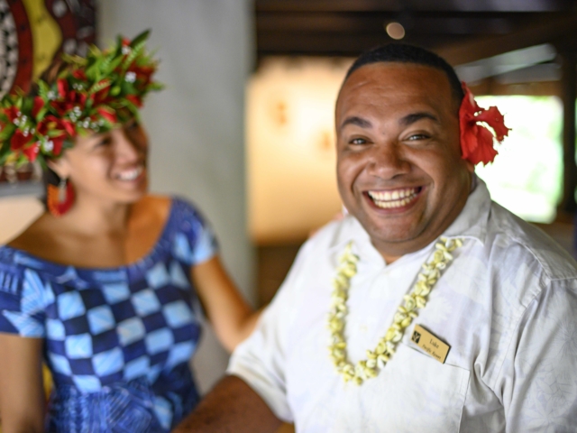 A happy full-faced smiling Resort attendant showcasing a beautiful red hibiscus flower behind his ear that symbolizes love and happiness